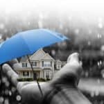 Hand holding a house with a blue umbrella in rain for waterproofing.