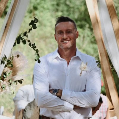 Smiling man in white shirt at outdoor event, greenery background.